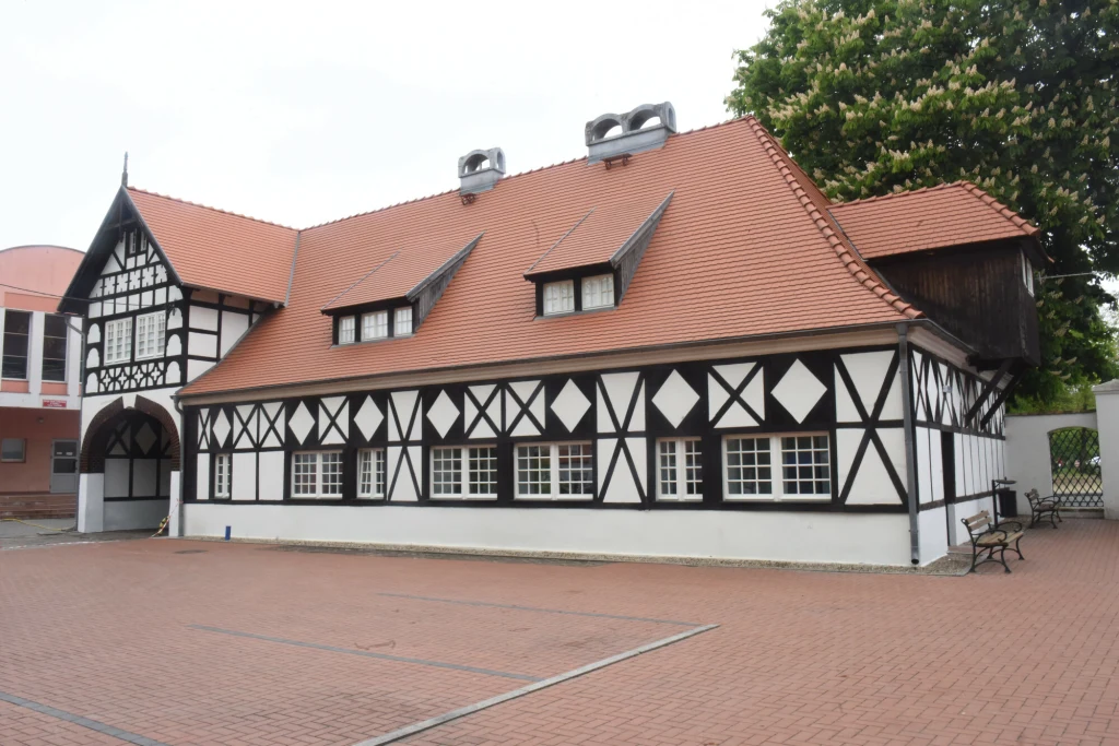 Budynek o konstrukcji szachulcowej z białymi ścianami i czarnymi belkami, dach kryty czerwoną dachówką, prostokątne okna rozmieszczone równomiernie, duży plac wyłożony czerwoną kostką brukową na pierwszym planie, w tle drzewa oraz fragment innych zabud...