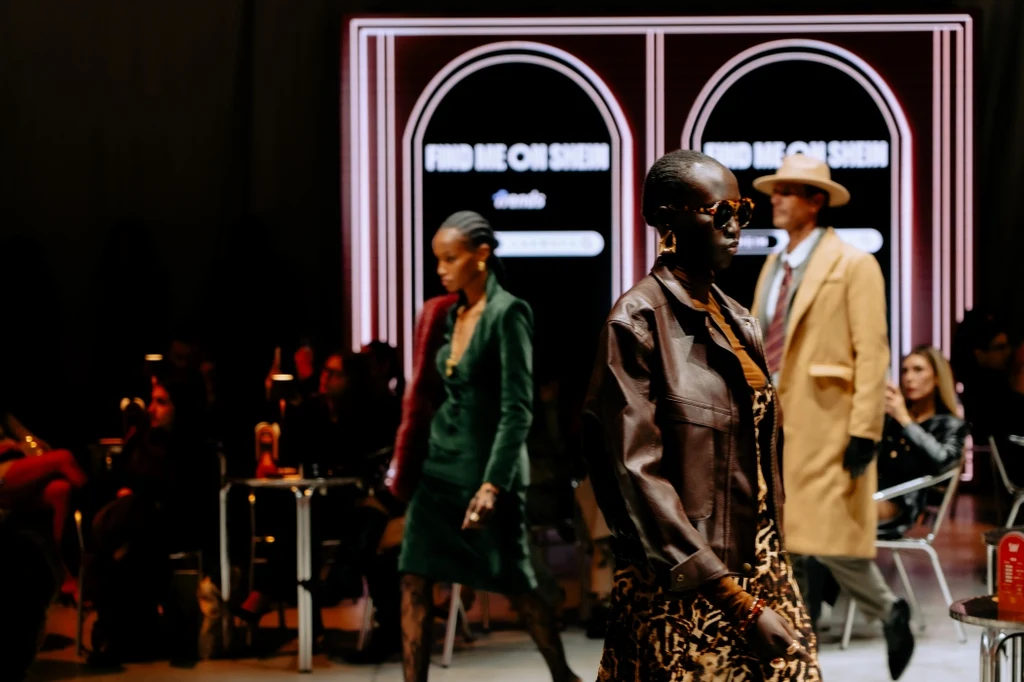 Pokaz mody z udziałem modeli noszących eleganckie ubrania, w tle widoczne światła i publiczność obserwująca wydarzenie, a na ścianie elektroniczny napis promocyjny.