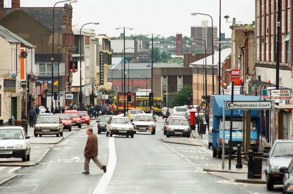 Ulica w Wielkiej Brytanii w latach 90. Ruchliwa ulica miejska otoczona budynkami, na której porusza się wiele samochodów, a przez przejście dla pieszych przechodzi starszy mężczyzna. Po prawej widoczna wiata przystankowa z napisem Magistrates Court, w tle zabudowa miejska i wysokie budynki.