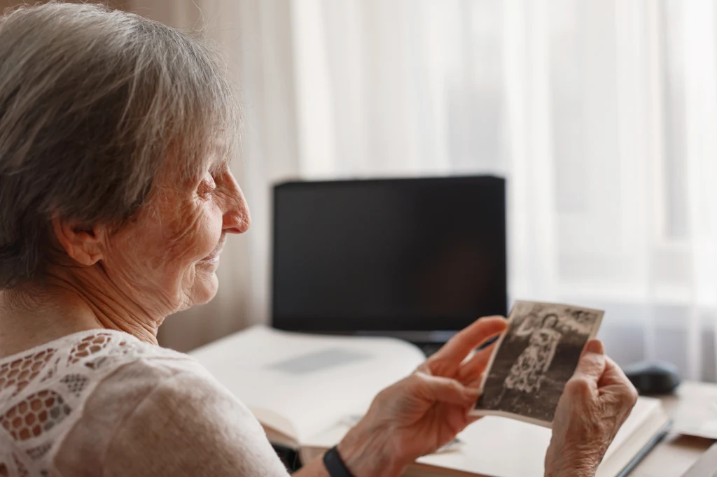 Starsza kobieta w białej bluzce trzyma czarno-białą fotografię, patrząc na nią z nostalgią. W tle znajduje się biurko z zamkniętym laptopem i jasnym światłem wpadającym przez zasłony.
