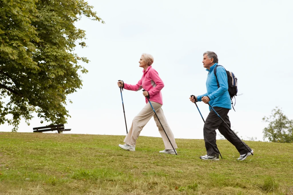 Starsza kobieta i mężczyzna spacerują po zielonym trawniku, wyposażeni w kije do nordic walkingu, ubrani w sportowe stroje, w tle drzewo i ławka.