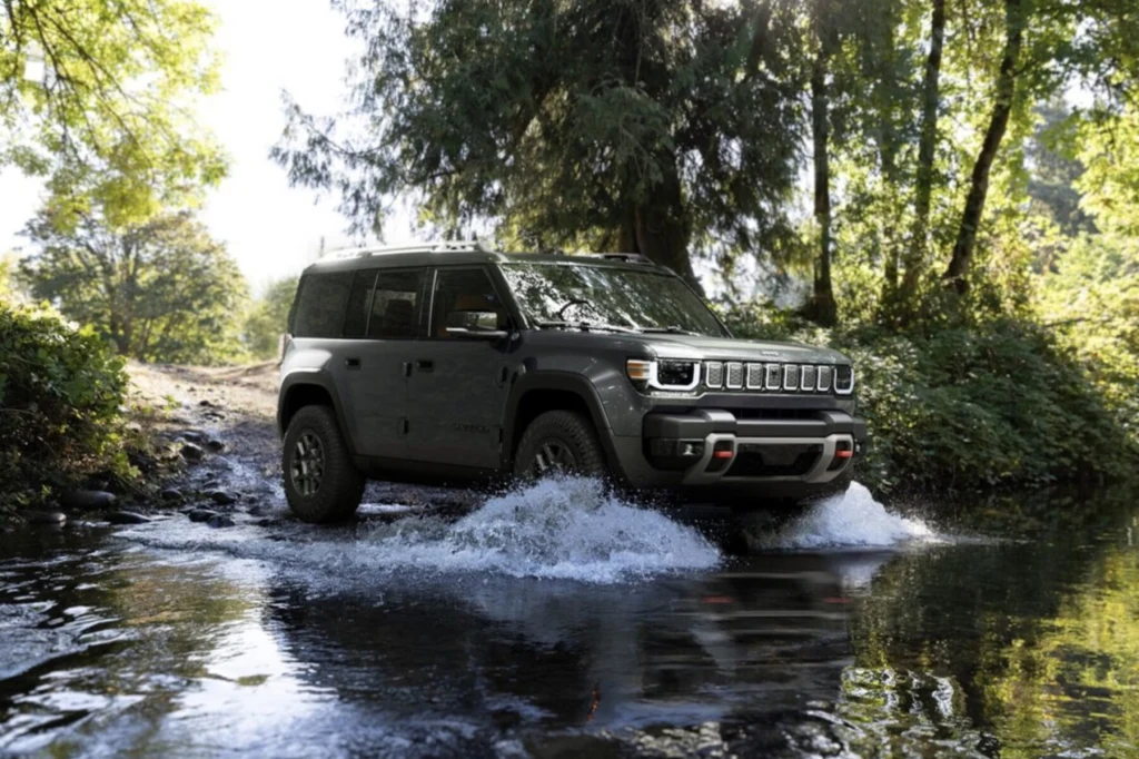 Samochód terenowy w ciemnym kolorze pokonuje płytką rzekę, wokół roślinność leśna i silne światło słoneczne przebijające przez korony drzew.