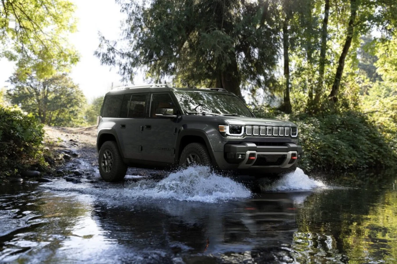 Samochód terenowy w ciemnym kolorze pokonuje płytką rzekę, wokół roślinność leśna i silne światło słoneczne przebijające przez korony drzew.
