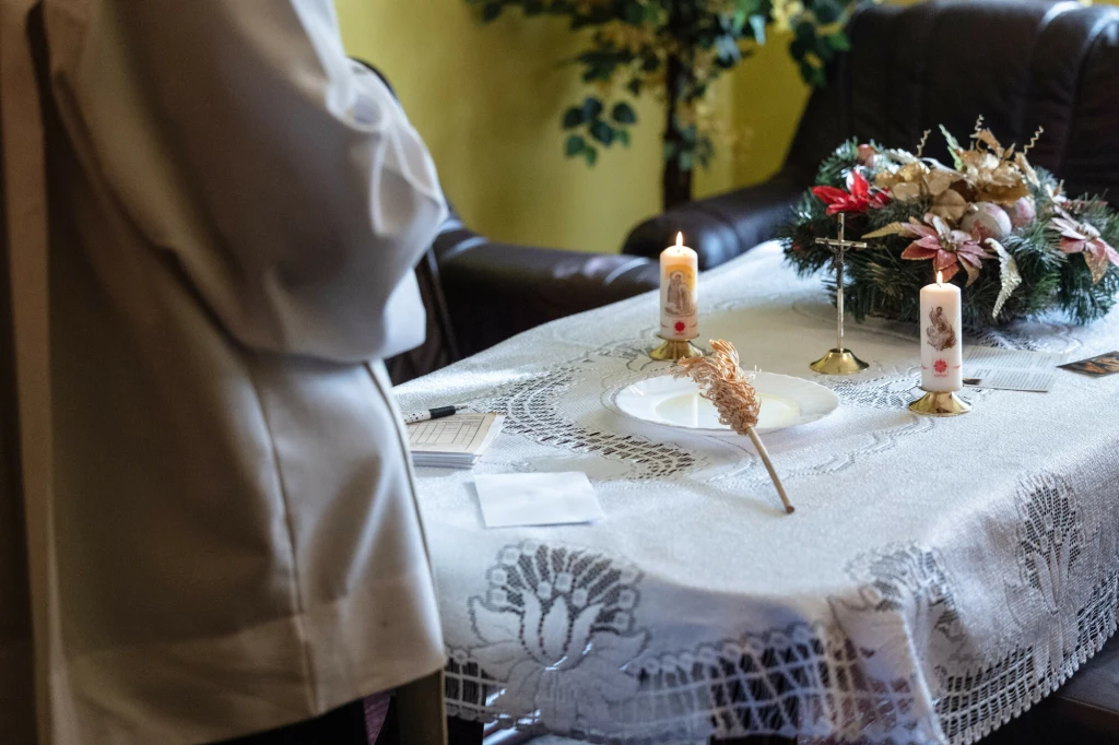 Wiele parafii przygotowuje się do kolędy. Tym razem wiernych czekają zmiany Stół nakryty białym obrusem z haftem, na którym stoją zapalone świece, gałązka do kropienia wodą święconą oraz stroik świąteczny. Po lewej stronie fragment osoby w białej szacie. W tle kanapa oraz roślina ozdobna.