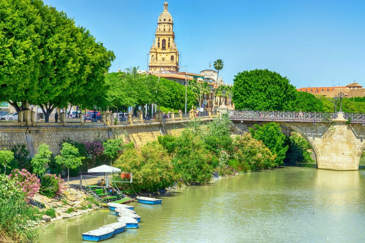 Rzeka przepływająca obok zielonych drzew, u brzegu zacumowane łódki, na drugim planie historyczny most z kamienia, nad którym góruje efektowna wieża zabytkowego budynku wśród bujnej zieleni.