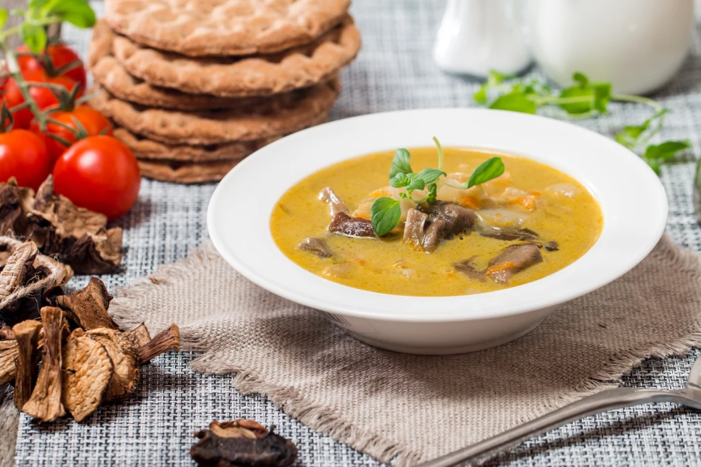 Zupa grzybowa to idealny wybór na czas Adwentu Biała miska z zupą grzybową, udekorowana świeżą natką pietruszki. Obok znajdują się suszone grzyby, kilka pomidorów oraz chrupiące pieczywo chrupkie ułożone w stos. Całość ustawiona na lnianej serwetce i jasnym tle stołu.