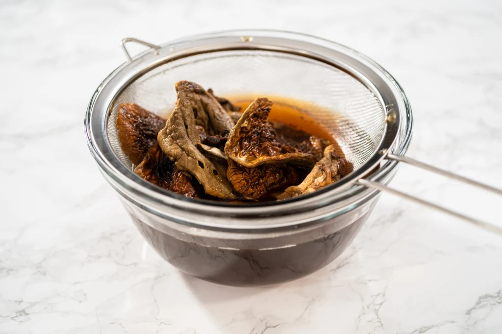 Przed moczeniem grzybów, koniecznie połam je na mniejsze kawałki Namoczone suszone grzyby oddzielone od aromatycznego wywaru za pomocą metalowego sitka i szklanej miski, całość ustawiona na jasnym, marmurowym blacie.