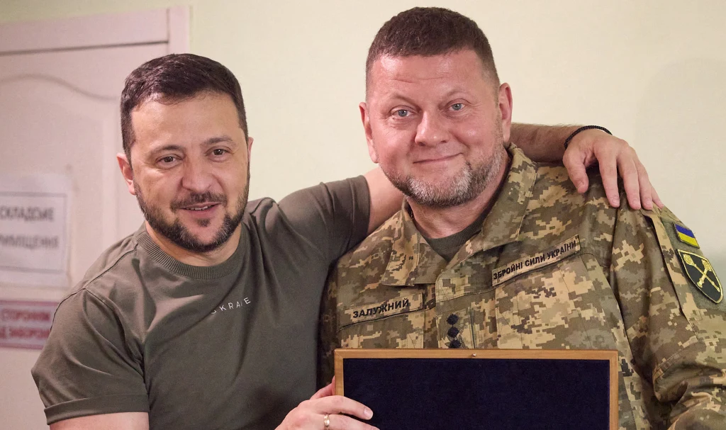 Dwóch mężczyzn stojących obok siebie, jeden w wojskowym mundurze z odznakami, drugi w zielonej koszulce, obejmują się, obaj uśmiechnięci, w tle jasne wnętrze i zamknięte drzwi.