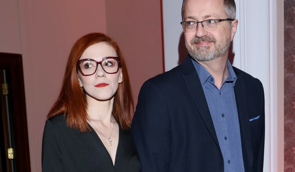 Przemysław i Gabriela Borkowscy Kobieta z długimi rudymi włosami, w dużych okularach oraz mężczyzna w marynarce i okularach stoją razem w eleganckim pomieszczeniu, oboje patrzą w różnych kierunkach.