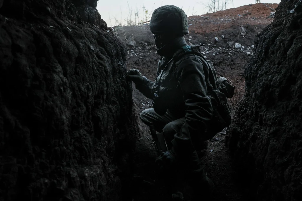Sylwetka żołnierza w umundurowaniu i hełmie, kucającego w ciemnym okopie na tle ziemistego, surowego terenu. Twarz niewidoczna przez cień, atmosfera napięcia i przygotowania do walki.