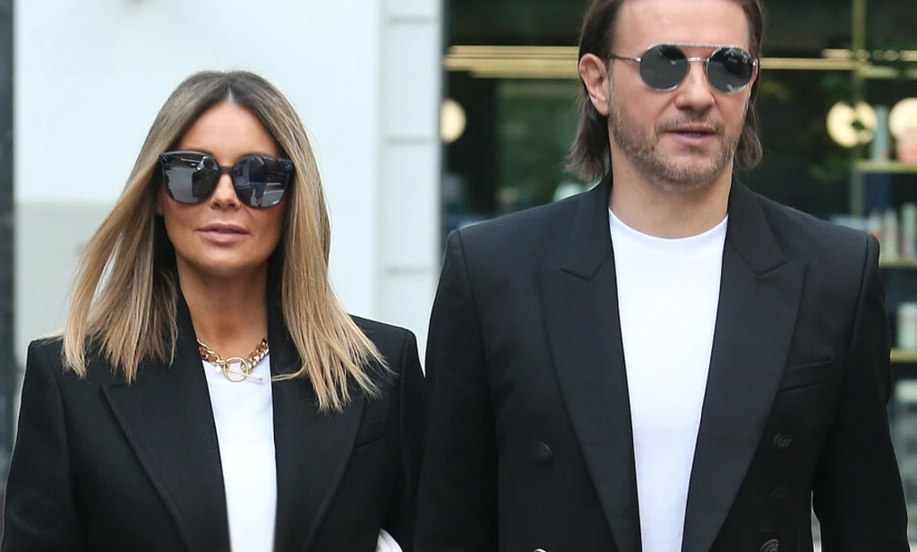 Para eleganckich osób w ciemnych okularach i czarnych marynarkach, spacerująca razem ulicą w przestrzeni miejskiej. Oboje mają poważne wyrazy twarzy i białe koszulki pod marynarkami.