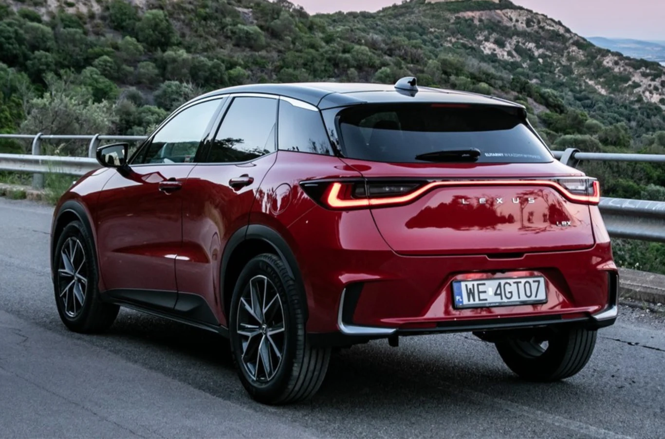 Nowoczesny, czerwony samochód SUV zaparkowany na asfaltowej drodze, z widoczną tablicą rejestracyjną i efektownymi światłami tylnymi, otoczony zielonymi wzgórzami oraz poręczą przy drodze.