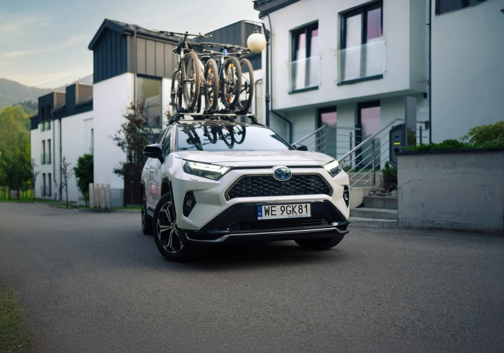 Biały samochód osobowy Toyota zaparkowany na spokojnej ulicy przed nowoczesnym budynkiem mieszkalnym, na dachu pojazdu zamocowane dwa rowery.