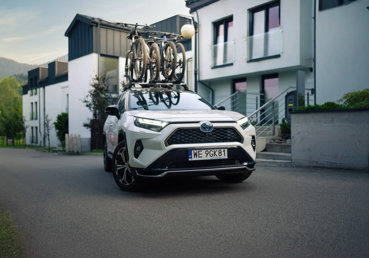 Biały samochód osobowy Toyota zaparkowany na spokojnej ulicy przed nowoczesnym budynkiem mieszkalnym, na dachu pojazdu zamocowane dwa rowery.