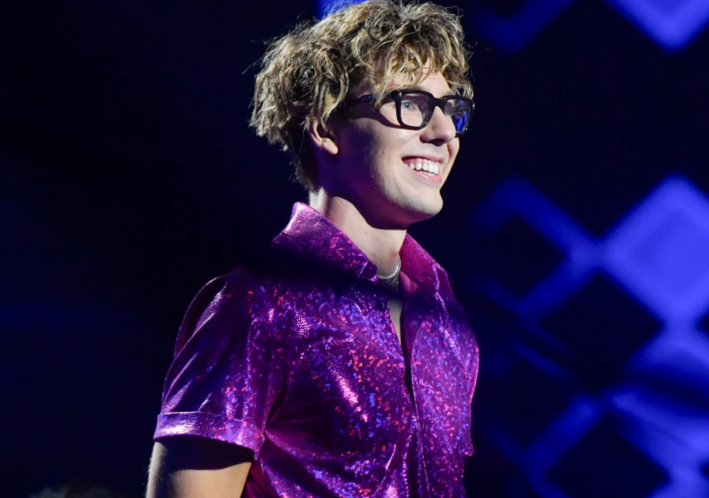 Mikołaj "Bagi" Bagiński Młody mężczyzna w okularach i błyszczącej, fioletowej koszuli uśmiecha się szeroko, stojąc na tle niebiesko-fioletowych świateł scenicznych.