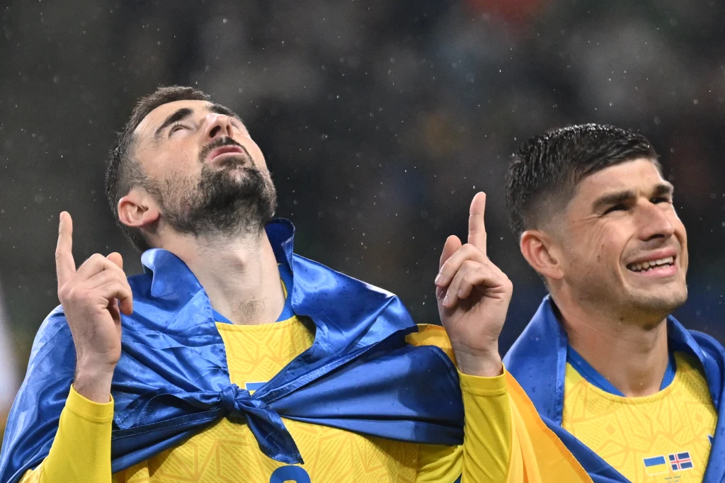 Dwóch piłkarzy w żółtych koszulkach oraz z niebieskimi flagami owiniętymi wokół ramion, obaj z uniesionymi palcami wskazującymi w górę, jeden z nich patrzy w niebo, obok siebie wyrażają radość i wzruszenie.