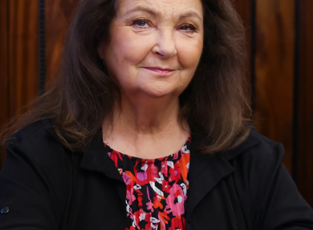 Anna DYmna odpowiada za organizację Festiwalu Zaczarowanej Piosenki Kobieta o długich, ciemnych włosach, ubrana w czarną marynarkę oraz kolorową sukienkę w czerwono-różowy wzór, siedzi na tle drewnianej ściany i patrzy z lekkim uśmiechem w stronę obiektywu.