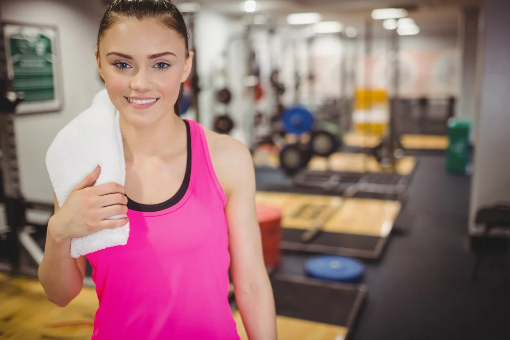Młoda kobieta w różowym topie sportowym z białym ręcznikiem na ramieniu stoi na tle sali treningowej wyposażonej w sprzęt fitness i podesty do ćwiczeń.
