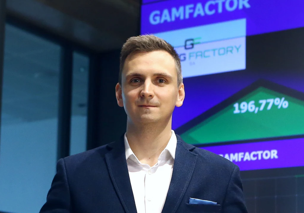 Mężczyzna w garniturze stoi na tle ekranu prezentującego wyniki firmy Gamfactory oraz wykres wzrostu wartości procentowej.