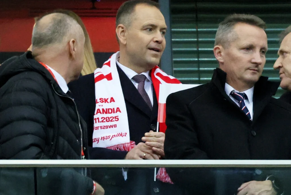 Grupa mężczyzn ubranych elegancko, stojących w loży stadionowej, jeden z nich ma na sobie biało-czerwony szalik z napisem związanym z meczem reprezentacji Polski.