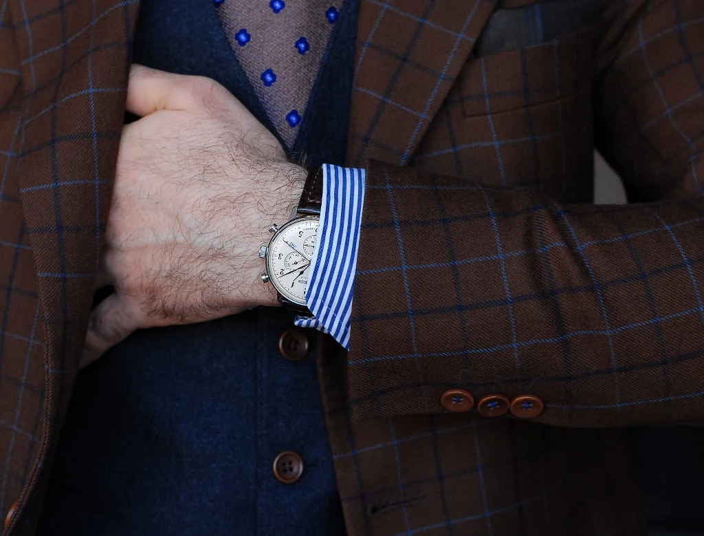 Marka Zeppelin Brązowa marynarka w kratę nałożona na niebieską kamizelkę i koszulę w paski, widoczny elegancki zegarek na nadgarstku i fragment krawata w niebieskie kropki.