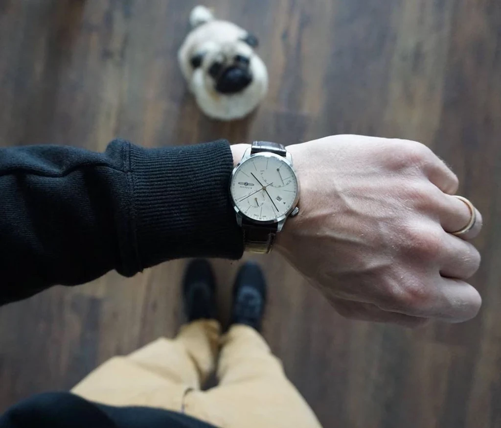 Zegarek Zeppelin jako idealny prezent Męska ręka z zegarkiem na nadgarstku wysunięta na pierwszy plan, w tle na podłodze stoi pies rasy mops i widoczne są nogi właściciela w jasnych spodniach oraz czarnych butach.
