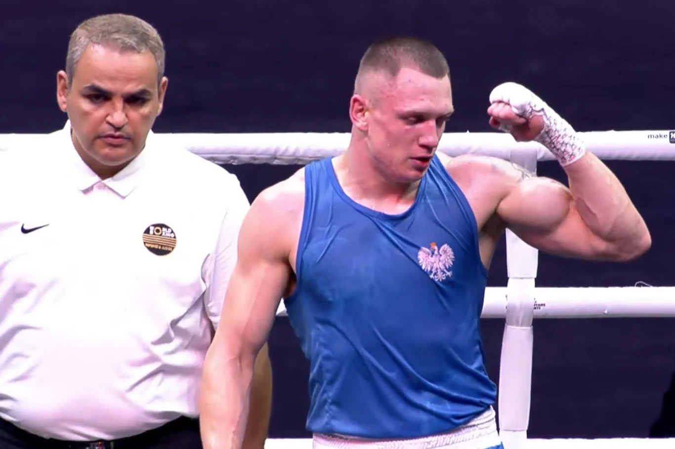 Bokser w niebieskiej koszulce z godłem Polski zaciska pięść w geście zwycięstwa, obok stoi sędzia w białej koszuli i obserwuje zawodnika, tło ciemne i neutralne, na pierwszym planie wyraźna emocja sportowa.