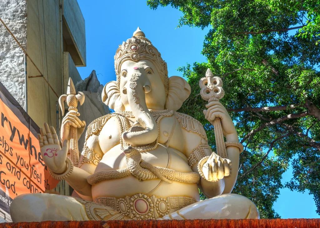 Ganeśa to jeden z najbardziej czczonych hinduskich bogów, charakteryzujący się głową słonia i ludzkim ciałem Monumentalna rzeźba Ganeszy, hinduskiego boga o głowie słonia, otoczona zielenią drzew oraz fragmentem nowoczesnego budynku. Postać wykonana z dużą dbałością o szczegóły, z licznymi ozdobami i w tradycyjnej pozie.