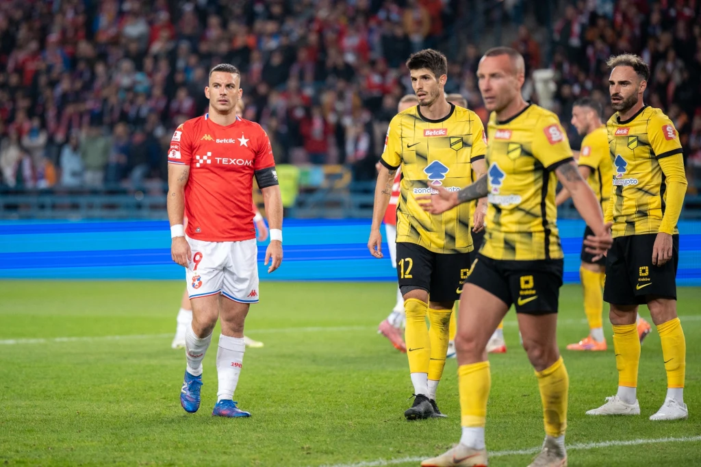 Piłkarze dwóch drużyn podczas meczu piłki nożnej na stadionie, jeden zawodnik w czerwonej koszulce, pozostali w żółto-czarnych strojach na tle licznie zgromadzonej publiczności.