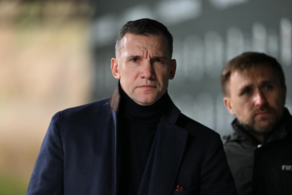 Andrij Szewczenko Mężczyzna w granatowym płaszczu i czarnym golfie patrzy poważnie przed siebie, w tle drugi mężczyzna ubrany w sportową kurtkę z napisem FIFA, lekko rozmyte tło.