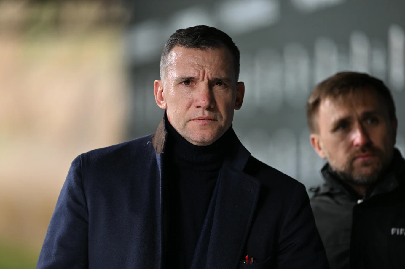 Mężczyzna w granatowym płaszczu i czarnym golfie patrzy poważnie przed siebie, w tle drugi mężczyzna ubrany w sportową kurtkę z napisem FIFA, lekko rozmyte tło.