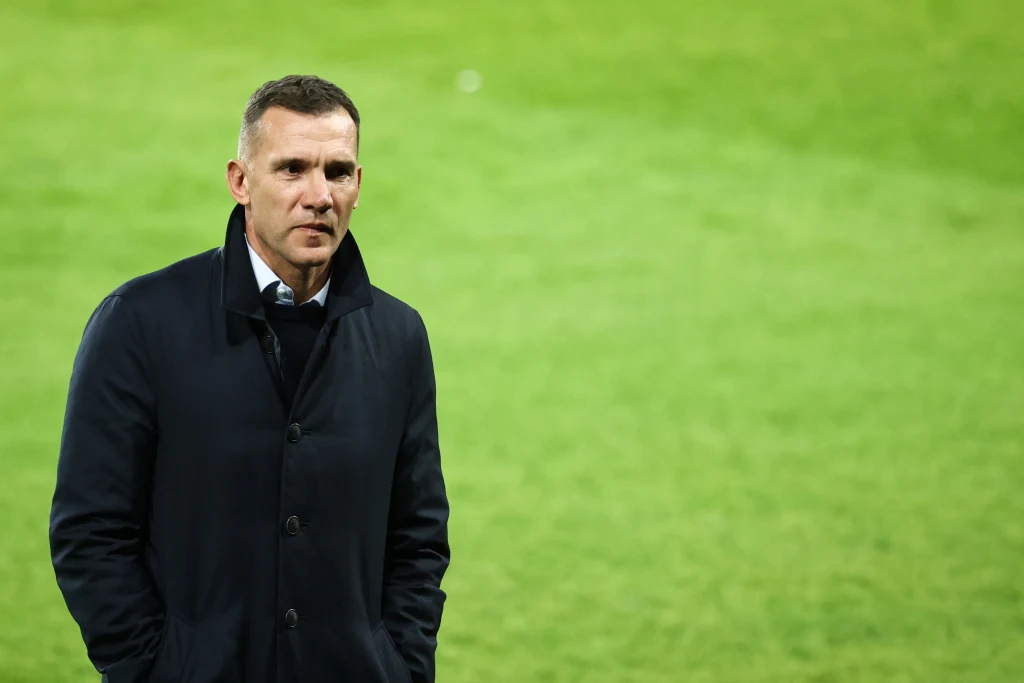 Mężczyzna w ciemnym płaszczu stoi na tle jasnozielonej murawy, z rękami w kieszeniach, patrzy spokojnie przed siebie.