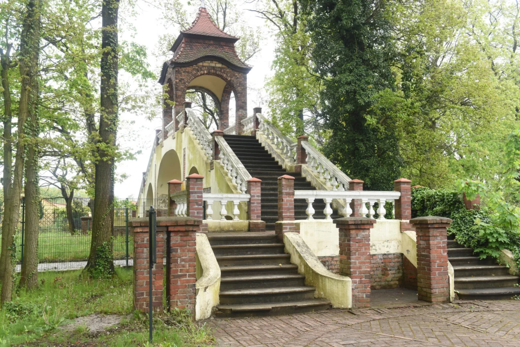 Kamienna ozdobna brama schodowa z balustradą prowadzącą na podwyższenie, zwieńczona zadaszeniem, otoczona przez bujną zieleń i drzewa.