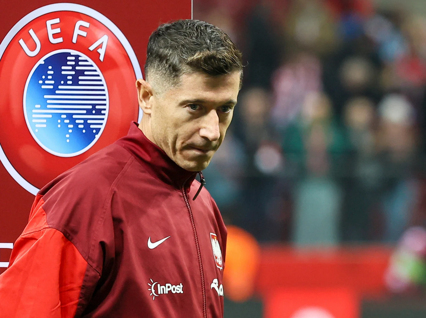 Polski piłkarz ubrany w czerwony dres reprezentacji stoi na tle dużego znaku UEFA, na stadionie podczas wydarzenia sportowego.