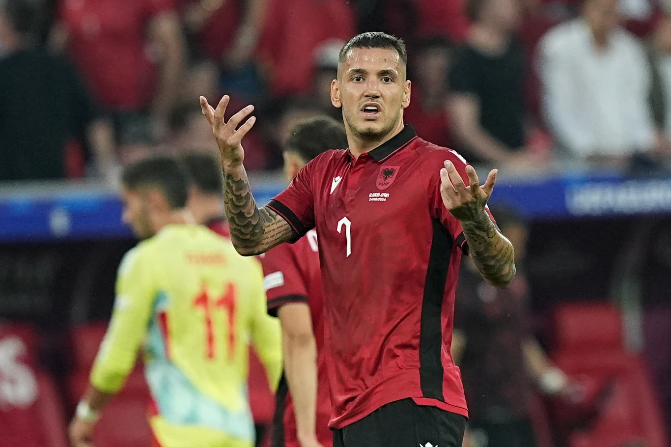 Rey Manaj, piłkarz reprezentacji Albanii Piłkarz ubrany w czerwoną koszulkę z numerem 7 i czarnymi dodatkami gestykuluje podczas meczu piłkarskiego, w tle widać innych zawodników oraz kibiców na trybunach.