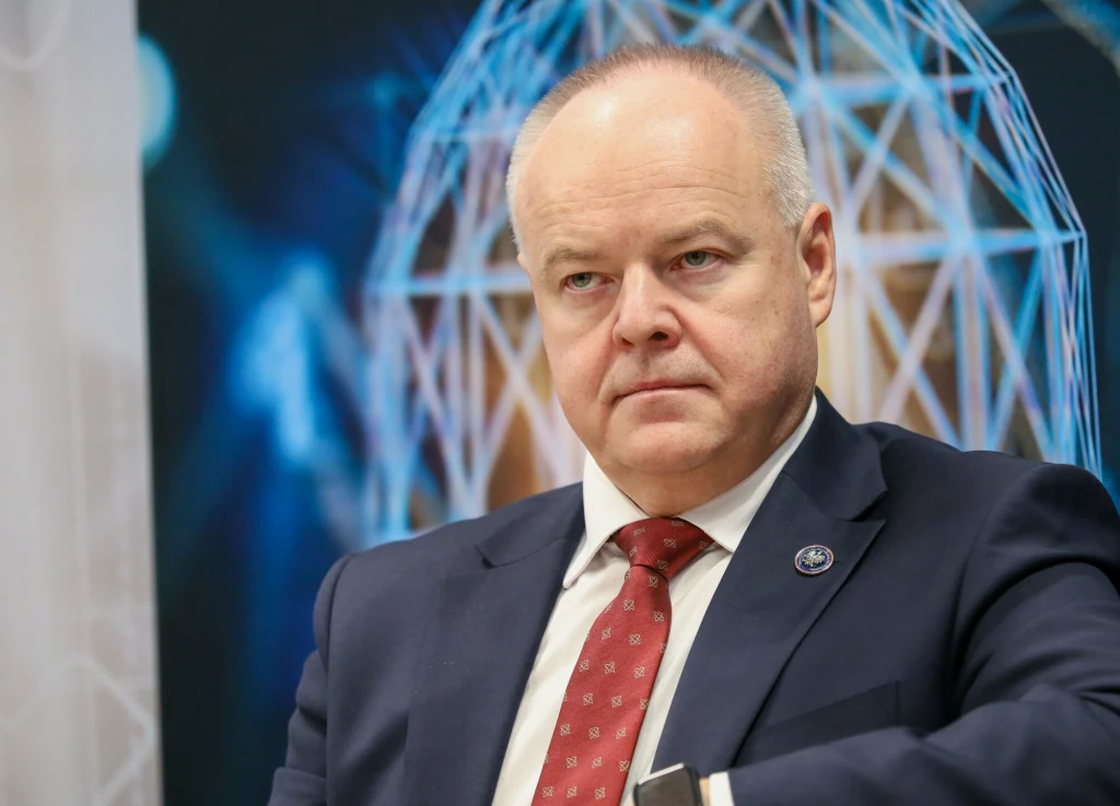 Mężczyzna w średnim wieku w garniturze i czerwonym krawacie siedzi na tle grafiki z geometryczną, świetlną strukturą przypominającą kopułę.