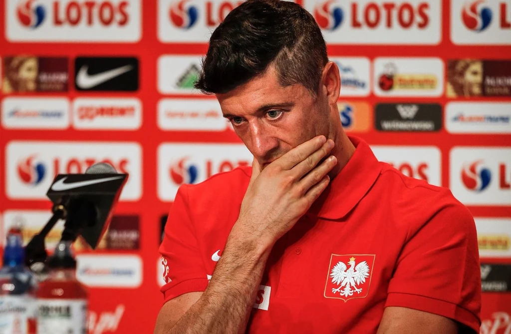 Robert Lewandowski Mężczyzna w czerwonej koszulce z godłem Polski siedzi przy stole podczas konferencji prasowej, w tle ścianka z logo sponsora, na twarzy zamyślony wyraz.