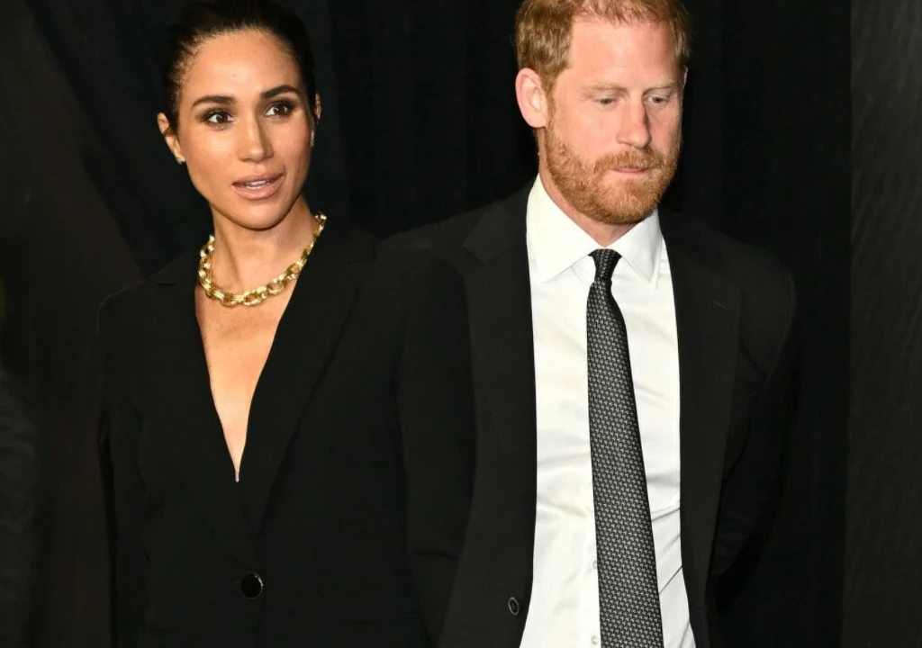 Meghan Markle i książę Harry Elegancka kobieta w czarnej marynarce i złotym naszyjniku stoi obok mężczyzny ubranego w ciemny garnitur z białą koszulą i krawatem, oboje patrzą w różne strony, tło ciemne, podkreślające ich stroje i wyraz twarzy.