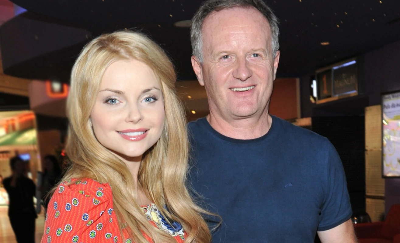 Dwoje uśmiechniętych ludzi pozujących razem, kobieta z blond włosami ubrana w kolorową sukienkę, mężczyzna w granatowej koszulce, w tle rozmyte wnętrze budynku z delikatnym oświetleniem.