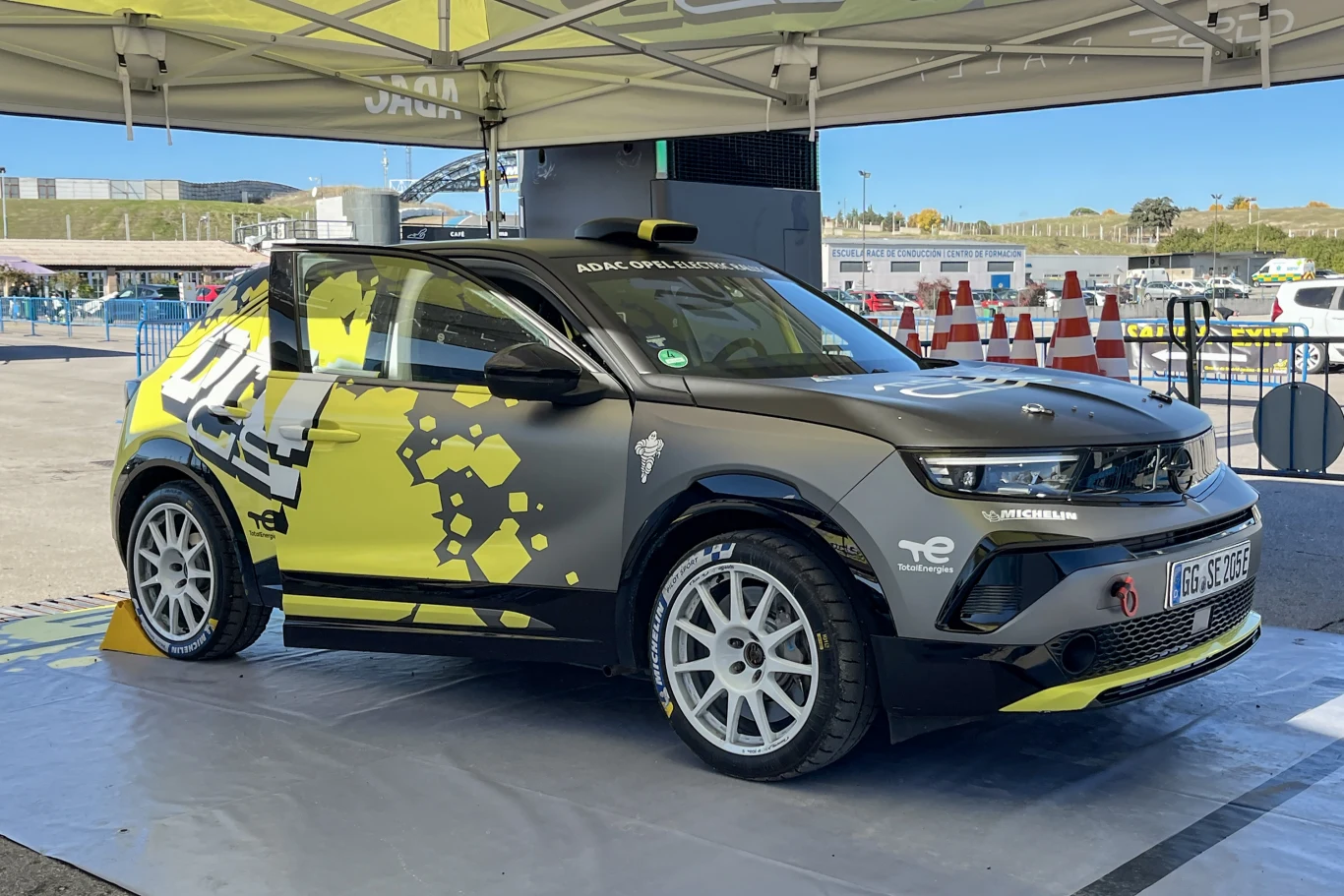 Sportowy samochód Opel Mokka Electric Rally w czarno-żółtym malowaniu wystawiony pod namiotem ekspozycyjnym, ustawiony na specjalnej platformie, w tle pachołki drogowe, fragment toru oraz otoczenie imprezy motoryzacyjnej.