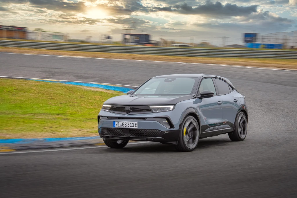 Szary samochód osobowy porusza się szybko po zakręcie asfaltowego toru wyścigowego, w tle widoczne są rozmyte trybuny oraz zachmurzone niebo z przebijającym światłem słonecznym.