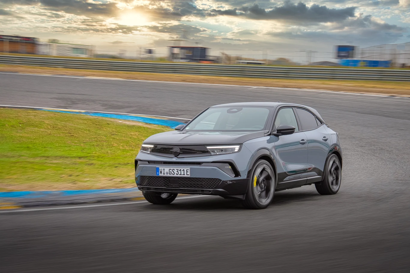 Szary samochód osobowy porusza się szybko po zakręcie asfaltowego toru wyścigowego, w tle widoczne są rozmyte trybuny oraz zachmurzone niebo z przebijającym światłem słonecznym.