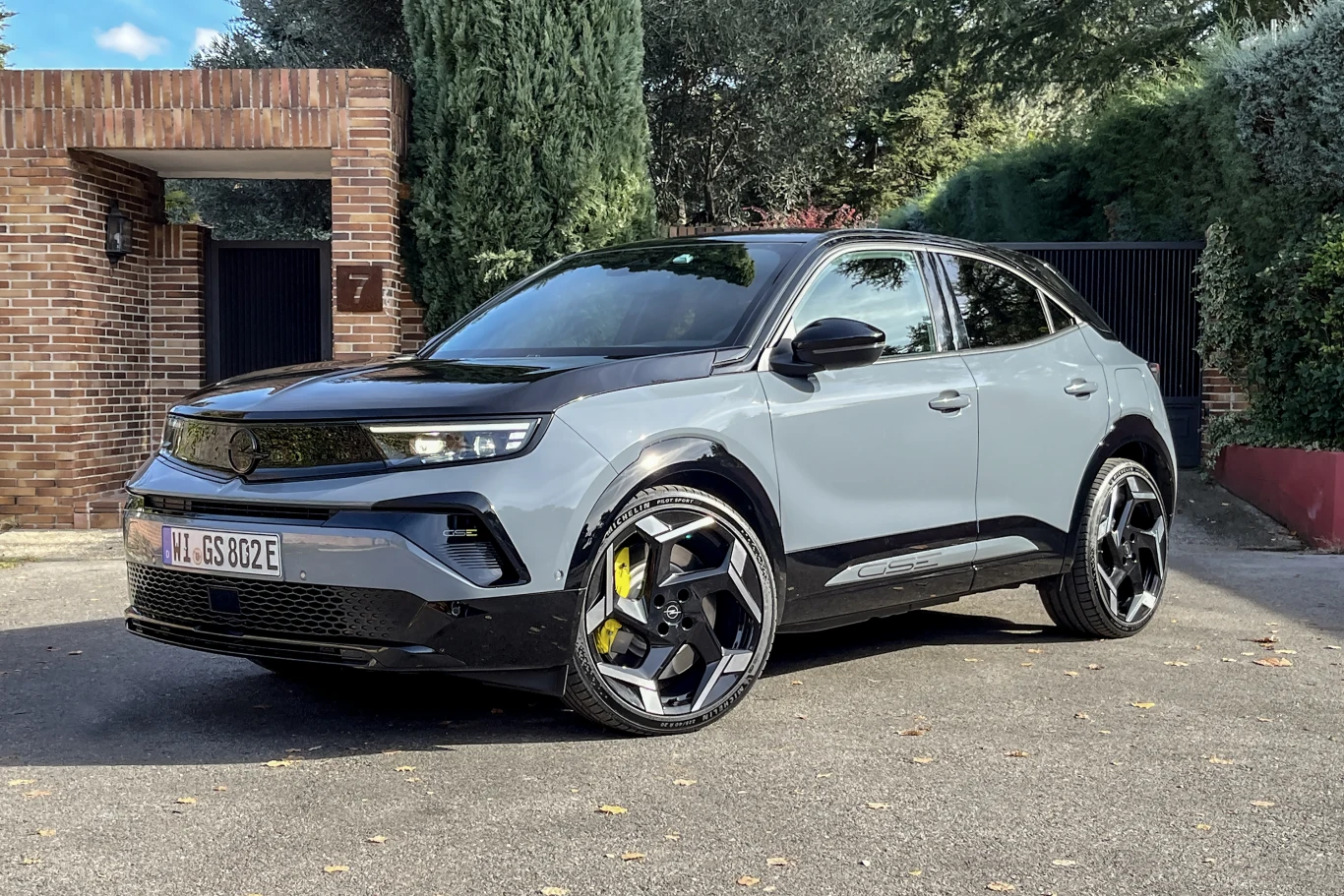 Nowoczesny samochód osobowy o sportowym wyglądzie w kolorze szaro-czarnym stoi zaparkowany na tle ceglanych murów i zieleni. Auto posiada duże, czarne felgi i żółte zaciski hamulcowe.