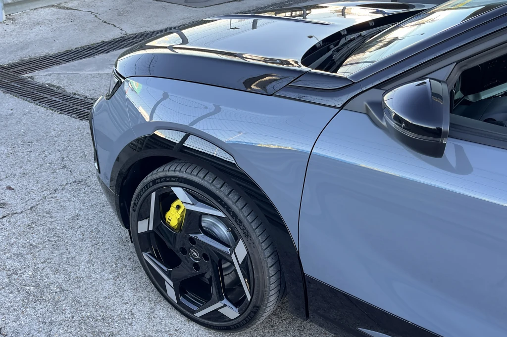 Szczegółowy widok przedniej części nowoczesnego samochodu w kolorze szarym z czarnymi akcentami, widoczne charakterystyczne felgi i żółty zacisk hamulcowy, całość stoi na betonowej nawierzchni.