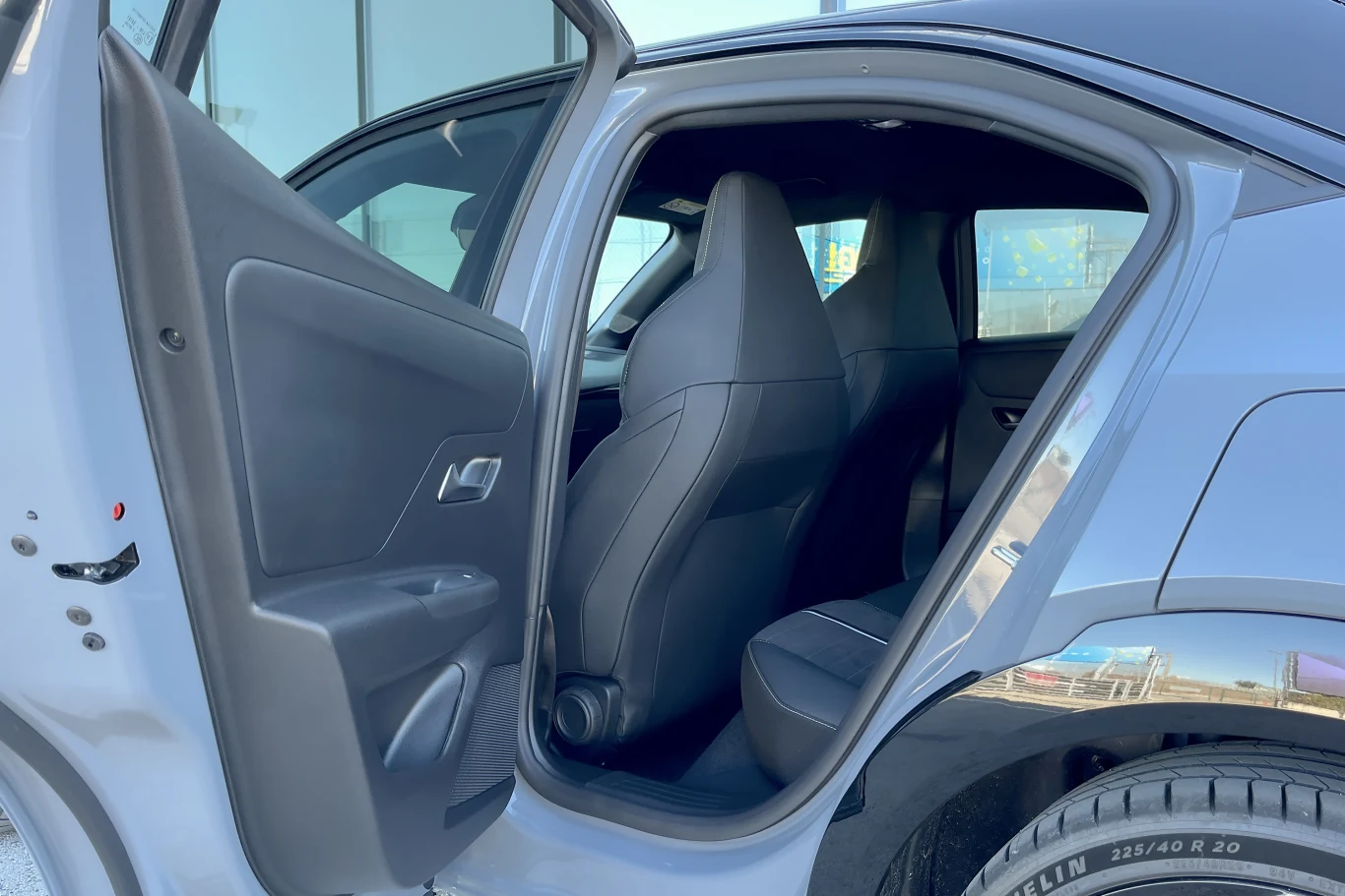 Wnętrze samochodu widoczne przez otwarte tylne drzwi, eksponujące tylną kanapę, czarne fotele oraz część przodu pojazdu z szybą i fragmentem opony.