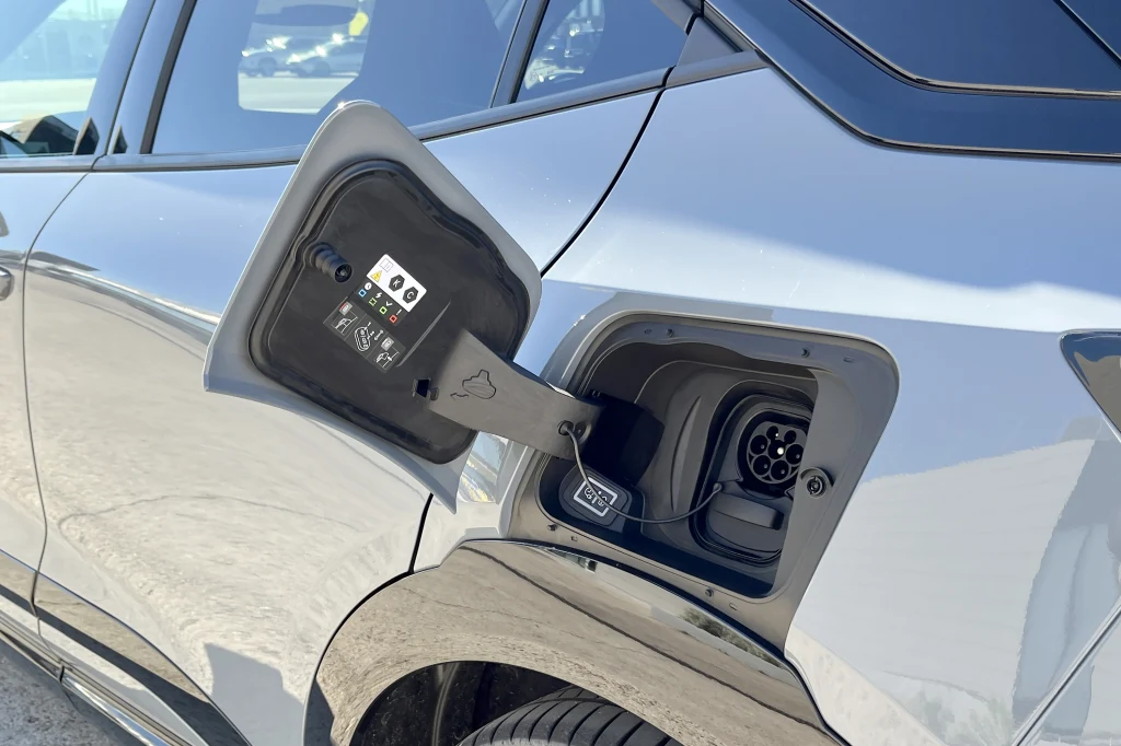 Otwarty wlew ładowania samochodu elektrycznego z widocznym gniazdem ładowania oraz instrukcjami na wewnętrznej stronie klapki, na tle srebrnego nadwozia auta.