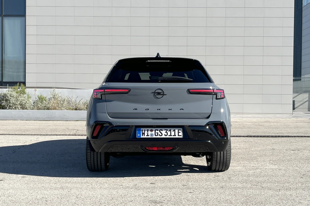 Szary samochód osobowy marki Opel Mokka sfotografowany od tyłu na tle nowoczesnego budynku z jasną elewacją. Auto stoi na wprost aparatu na jasnej nawierzchni, a na tablicy rejestracyjnej widoczny jest niemiecki numer.