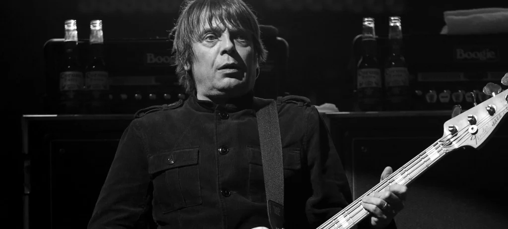 Mężczyzna na scenie grający na gitarze basowej, ubrany na czarno, w tle nagłośnienie i wzmacniacze, scena oświetlona reflektorami, atmosfera koncertowa.