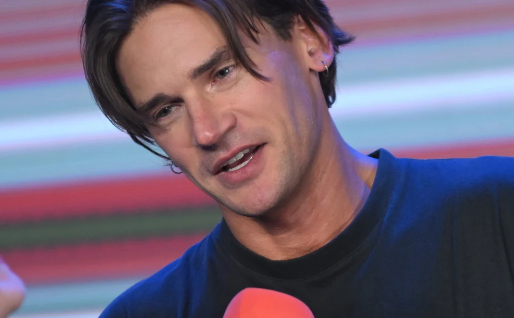 Michał Kot Młody mężczyzna z ciemnymi włosami rozmawia, trzymając czerwony mikrofon. W tle widoczne kolorowe, rozmyte pasy.