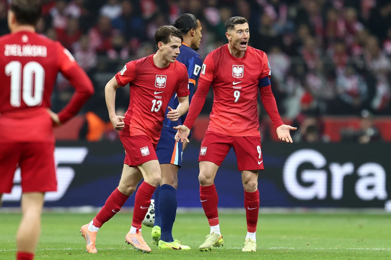 Dwóch piłkarzy w czerwonych strojach polskiej reprezentacji narodowej w emocjonującej dyskusji podczas meczu, wokół nich widoczni inni zawodnicy oraz kibice na trybunach.